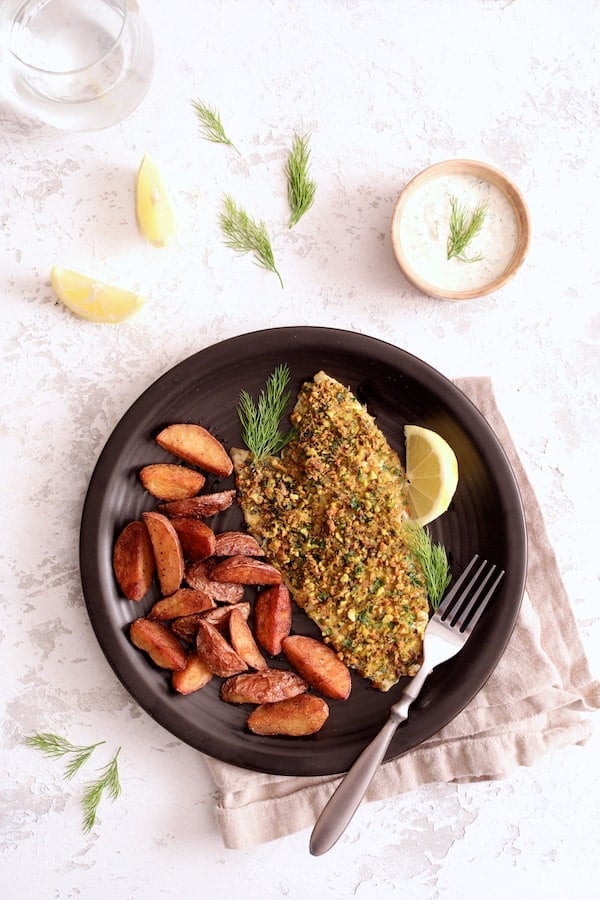 Photo of Pistachio-Crusted Fish with Lemon-Dill Aioli served with roasted potatoes garnished with lemon slices and dill sprigs on black plate.