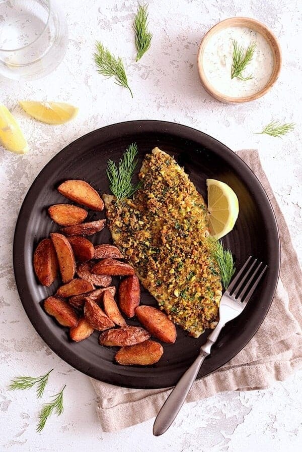One serving of Pistachio-Crusted Fish with Lemon-Dill Aioli with roasted potatoes on black dinner plate.