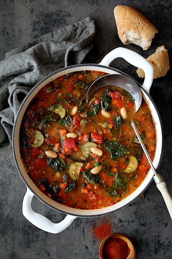 Photo of Smoky Spanish Vegetable and White Bean Soup with Kale in white Dutch oven with bread and paprika spilled on surface.