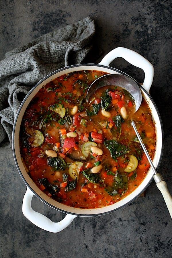 Photo of Smoky Spanish Vegetable and White Bean Soup with Kale in white Dutch oven on gray background with gray napkin and ladle.