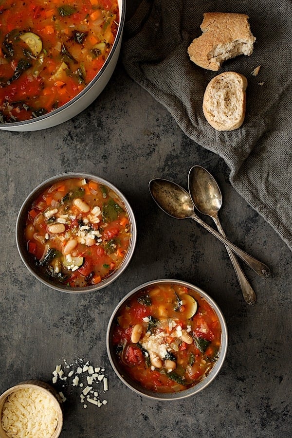 Photo of two servings of Smoky Spanish Vegetable and White Bean Soup with Kale with cheese and broken bread.