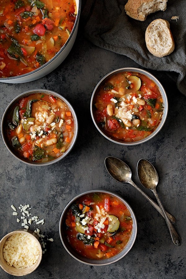 Photo of three bowls of Smoky Spanish Vegetable and White Bean Soup with Kale with broken bread and grated cheese.