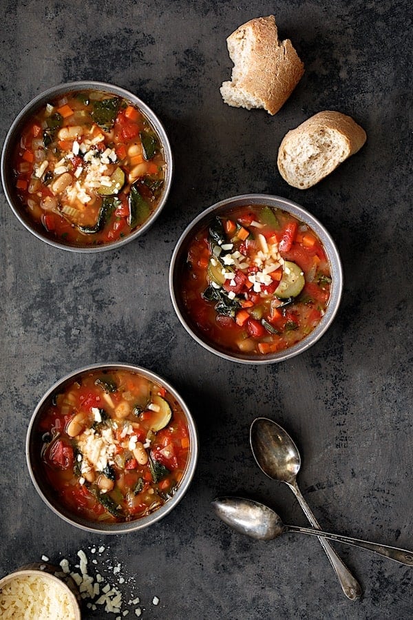 Photo of three bowls of Smoky Spanish Vegetable and White Bean Soup with Kale with broken bread and grated cheese.