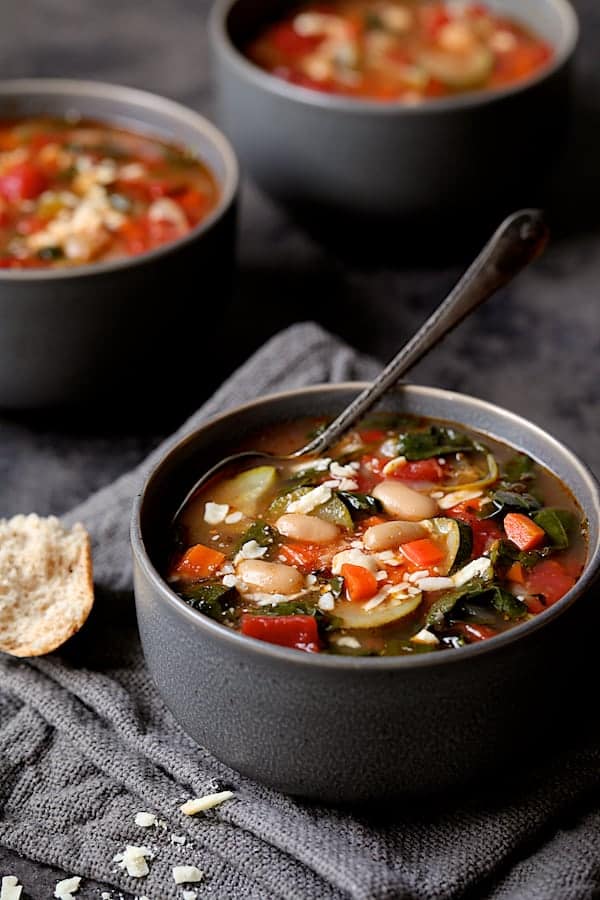 Close-up photo of Smoky Spanish Vegetable and White Bean Soup with Kale in gray bowl on gray napkin.