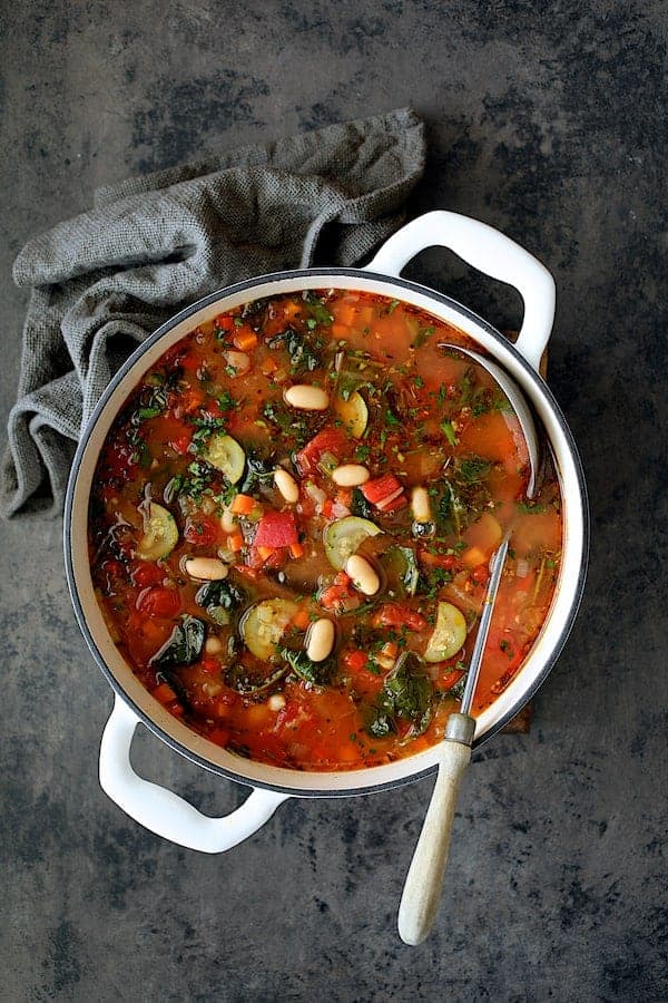 Photo of Smoky Spanish Vegetable and White Bean Soup with Kale in white Dutch oven on gray background with gray napkin and ladle.