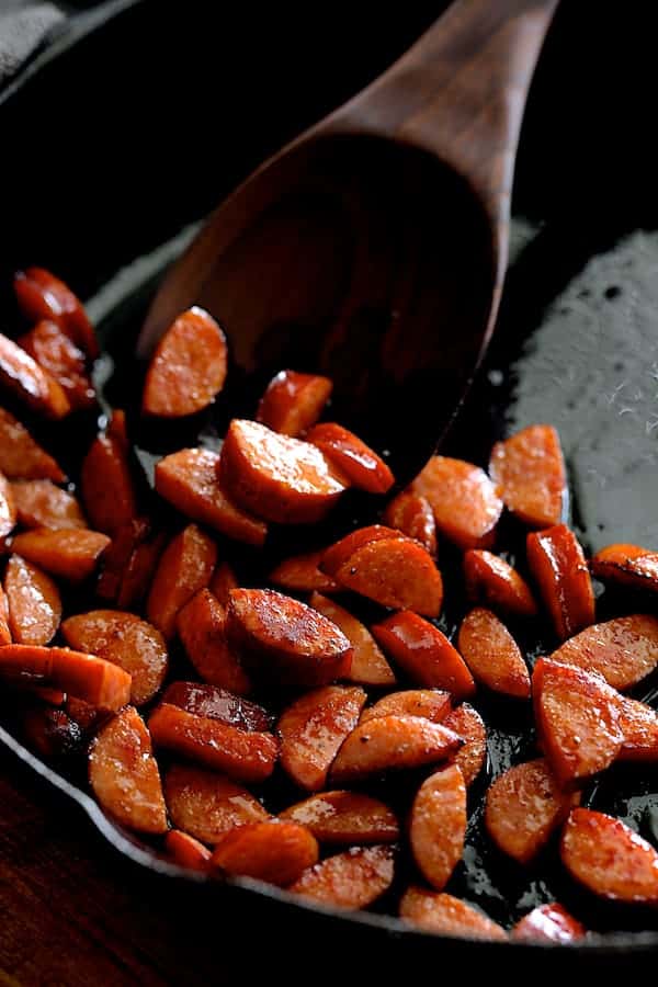 Photo of Andouille sausage being sauteed in cast iron skillet being stirred with wooden spoon