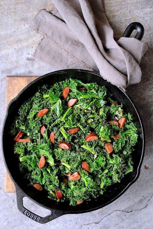 Spicy Sautéed Kale with Andouille Sausage - Overhead shot of dish in cast iron skillet on wooden trivet with gray towel