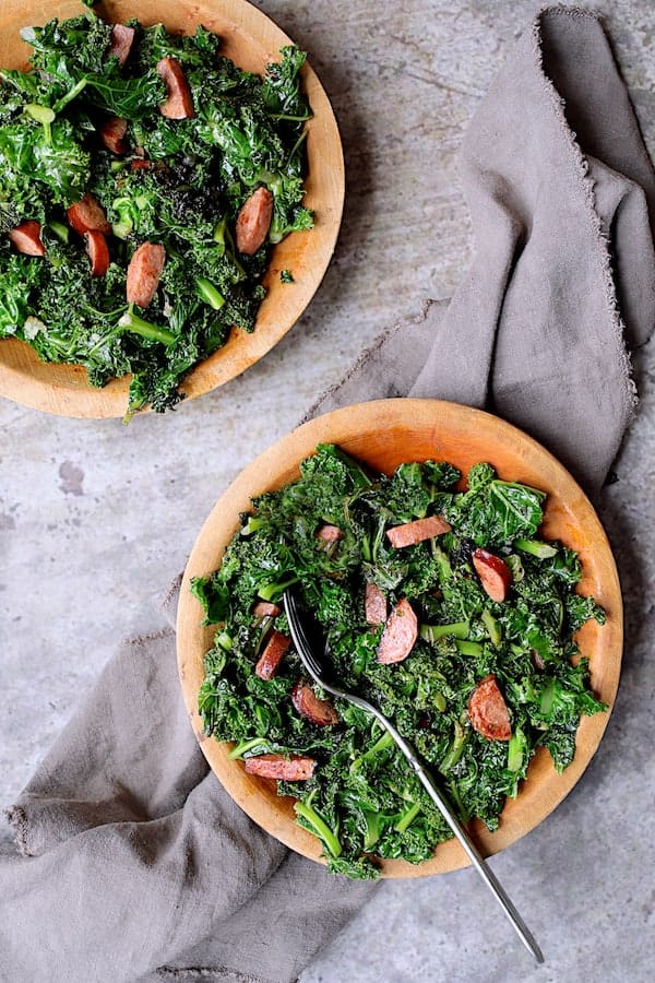 Spicy Sautéed Kale with Andouille Sausage - Overhead shot of dish on two wooden plates on gray background with gray napkin