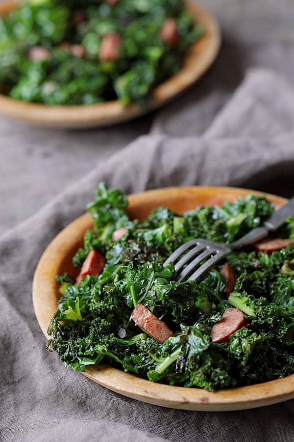Spicy Sautéed Kale with Andouille Sausage - Straight-on close-up shot of dish on wooden plate with fork stuck into sausage and kale on gray background