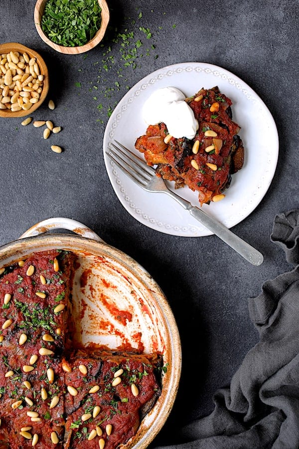 Photo of one piece of Turkish Eggplant Casserole - Imam Bayildi on a white plate with a dollop of yogurt the rest of the dish with wedge removed.