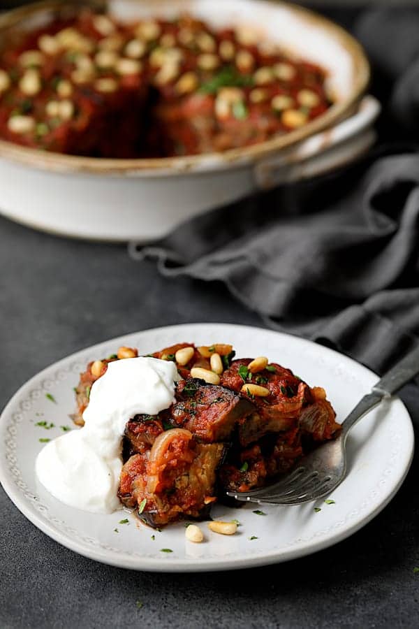 Close-up photo of one serving of Turkish Eggplant Casserole - Imam Bayildi on white plate with a dollop of yogurt with rest of the dish in the background.