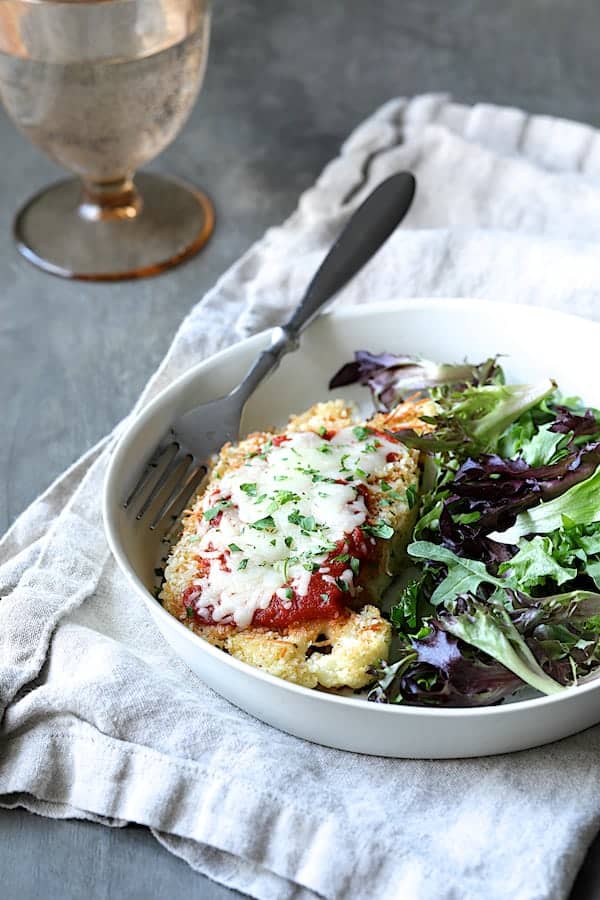 Close-up photo of Cauliflower Parmigiana in white dish with fresh baby greens.