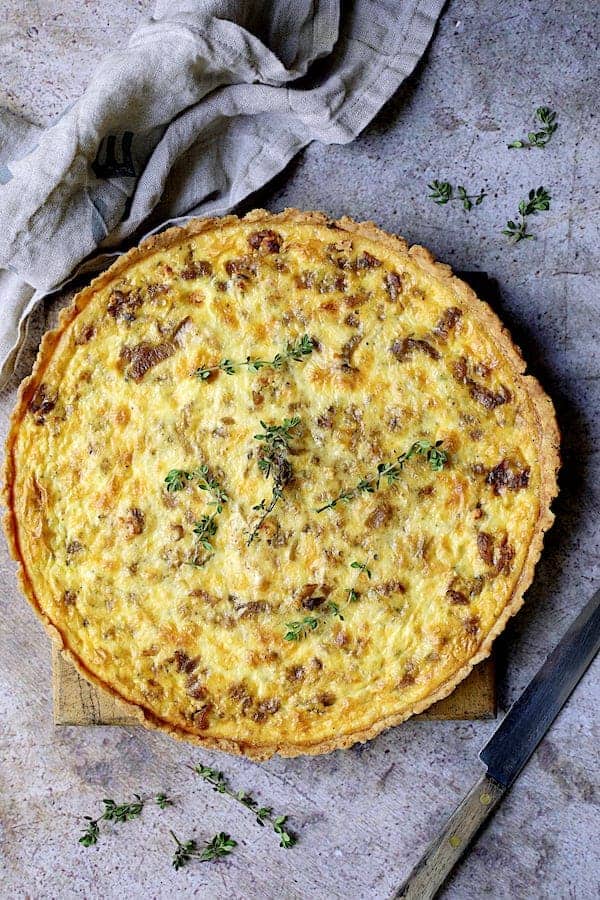 Photo of baked Caramelized Onion and Goat Cheese Tart on gray background with gray towel and knife.
