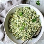 Cauliflower Artichoke Tabbouleh in white serving bowl with serving utensils garnished with fresh parsley.