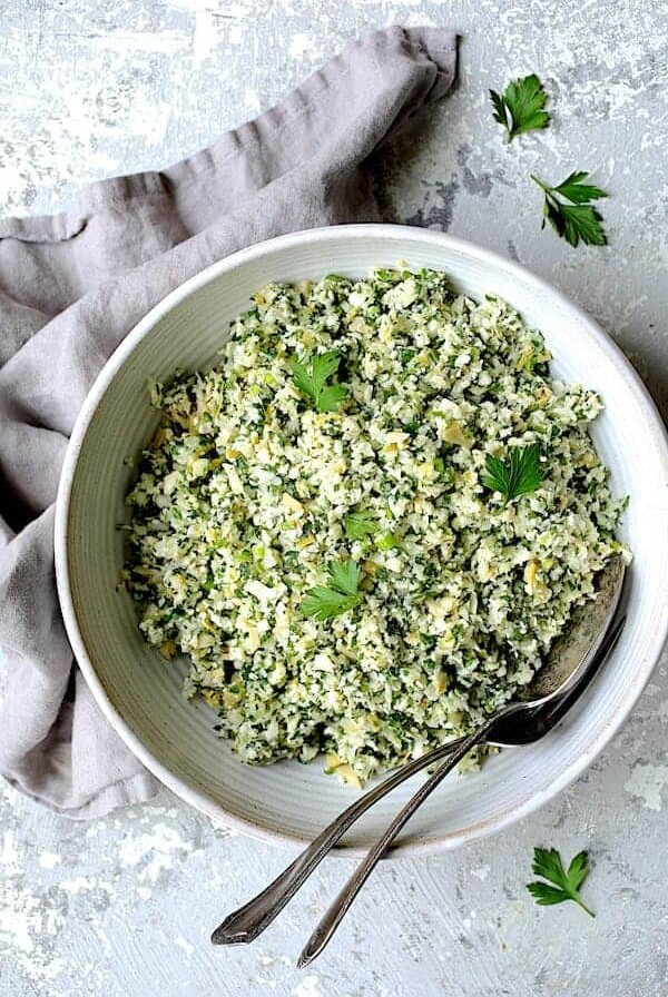 Cauliflower Artichoke Tabbouleh in white serving bowl with serving utensils garnished with fresh parsley.