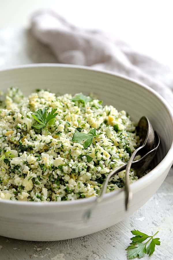Close-up photo of Cauliflower Artichoke Tabbouleh in white bowl with serving utensils garnished with parsley.