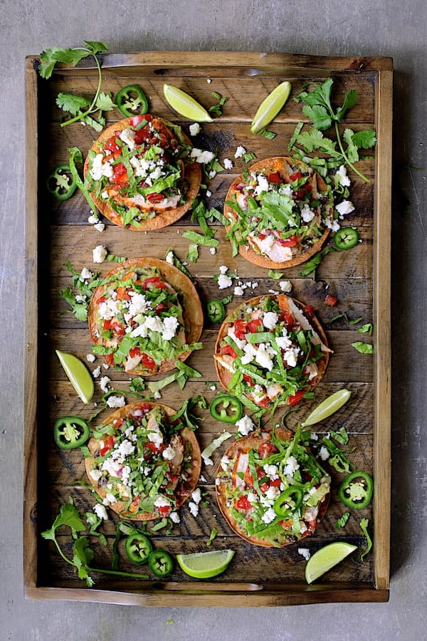 Photo of six Chicken Tostadas with Black Bean Guacamole and Salsa Fresca on wooden tray garnished with cilantro, lime wedges and jalapeno slices.