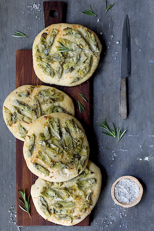 Photo of four individual loaves of Artichoke Focaccia with Rosemary on wooden breadboard with knife and garnished with fresh rosemary and coarse salt.