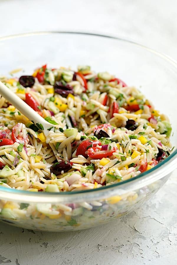 Photo of Greek Orzo Pasta Salad being mixed together with white spoon in glass bowl.