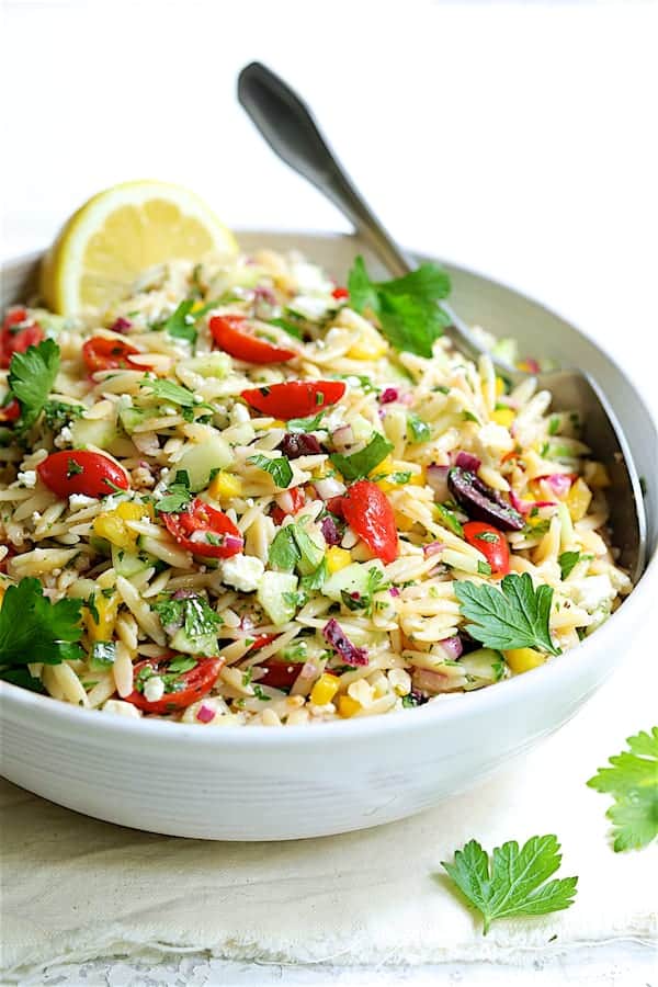 Close-up photo of Greek Orzo Pasta Salad in white serving bowl with serving spoon.