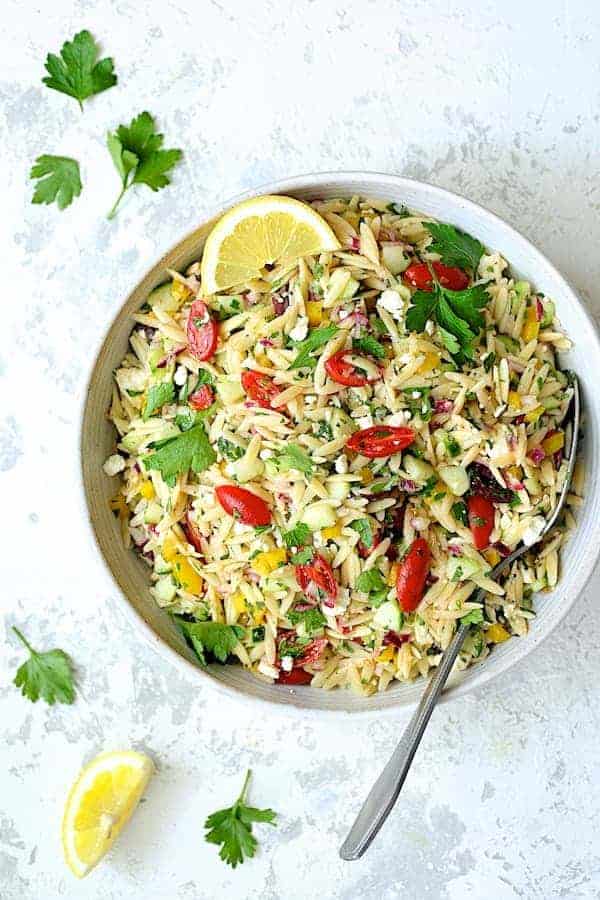 Photo of Greek Orzo Pasta Salad in white serving bowl on white distressed background with serving spoon, garnished with parsley and lemon.