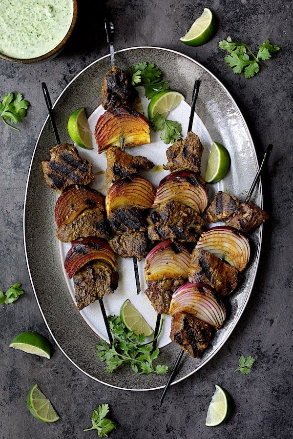 Indian-Style Beef Kabobs with Cilantro-Coconut Sauce - Overhead shot of cooked kabobs on gray-rimmed platter garnished with lime wedges and cilantro
