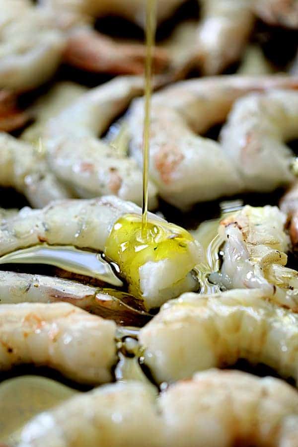 Photo of raw shrimp being drizzled with olive oil on baking sheet.