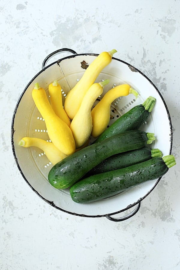 Photo of zucchini and yellow squash in white colander on white background.
