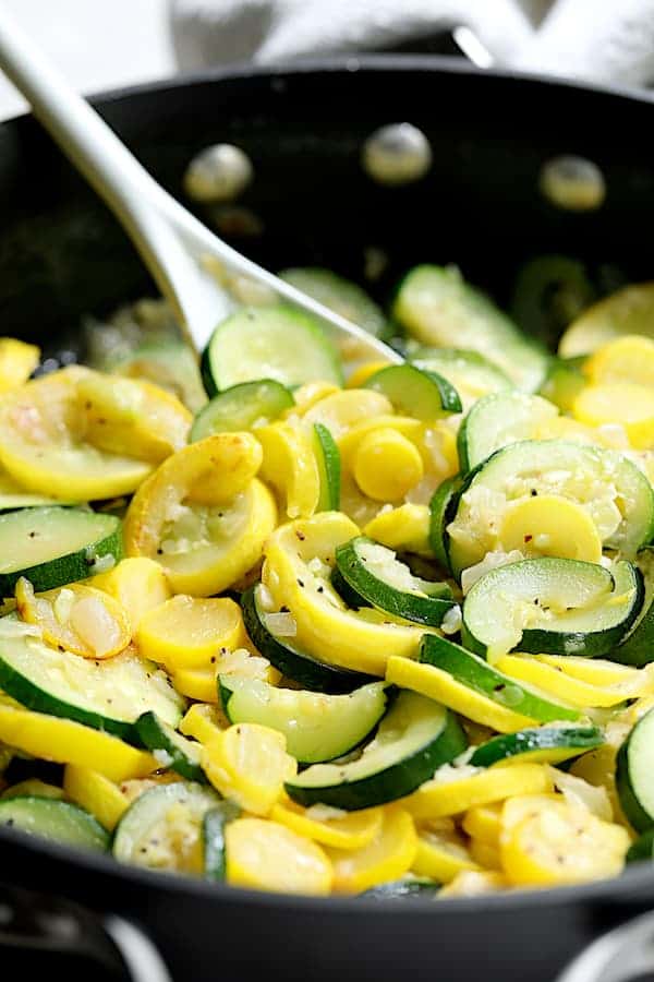 Photo of zucchini and yellow squash being sauteed in skillet with white spoon.