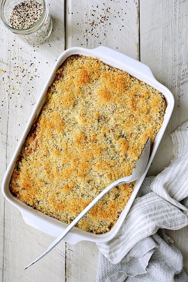 Photo of baked Zucchini, Yellow Squash and Quinoa Casserole on white wood background with white serving spoon and quinoa scattered about.