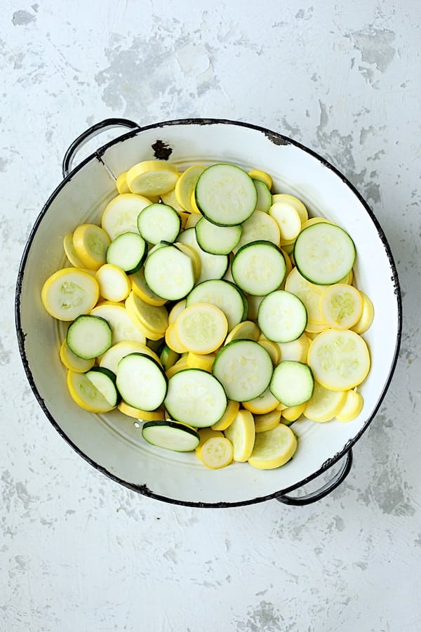 Photo of sliced zucchini and yellow squash in white colander on white background.