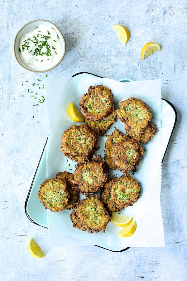 Photo of Zucchini Ricotta Fritters on light blue tray with yogurt garnished with chives and lemon.
