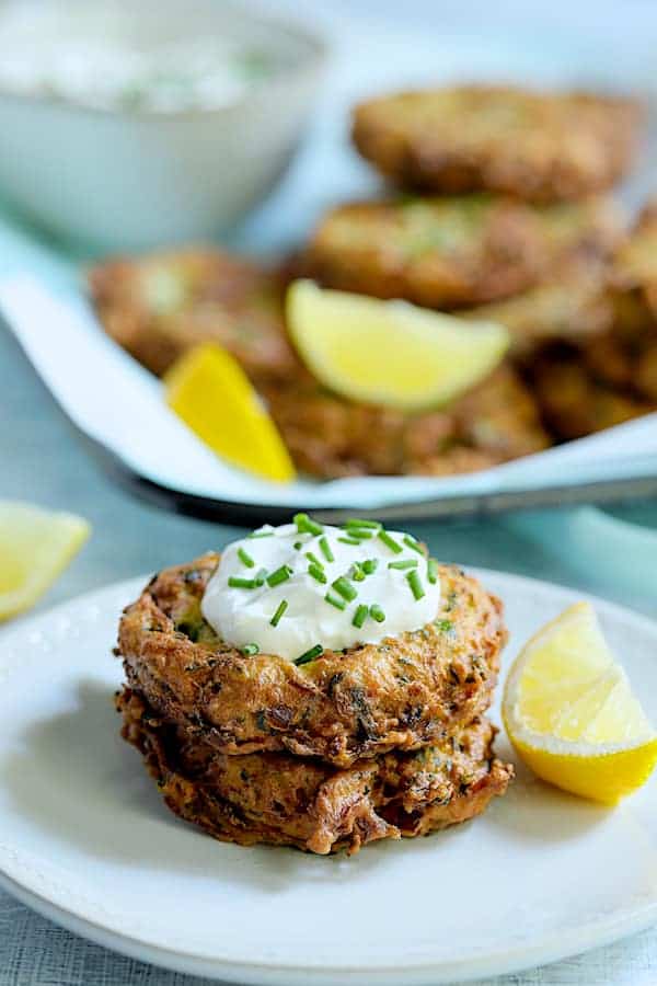Photo of two Zucchini Ricotta Fritters on white plate with yogurt on top garnished with fresh chives and lemon wedges.