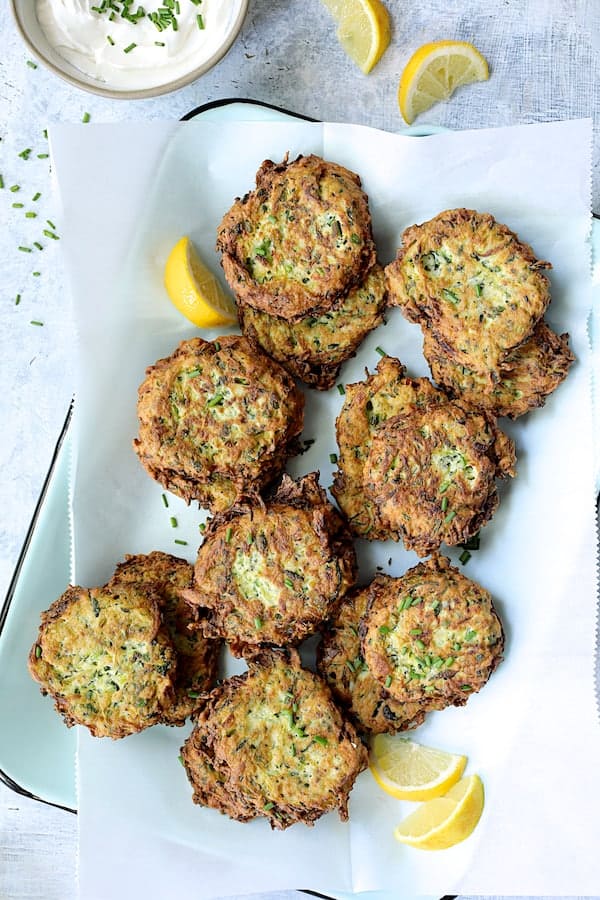 Photo of Zucchini Ricotta Fritters on parchment paper on light blue tray with lemon wedges.
