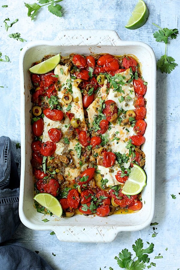Photo of Baked Veracruz-Style Fish (Pescado A La Veracruzana) in white baking dish on light blue background garnished with lime wedges and cilantro.