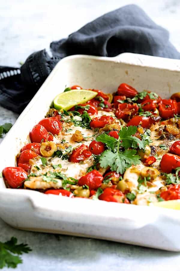 Close-up photo of Baked Veracruz-Style Fish (Pescado A La Veracruzana) in white baking dish with dark blue towel in the background.