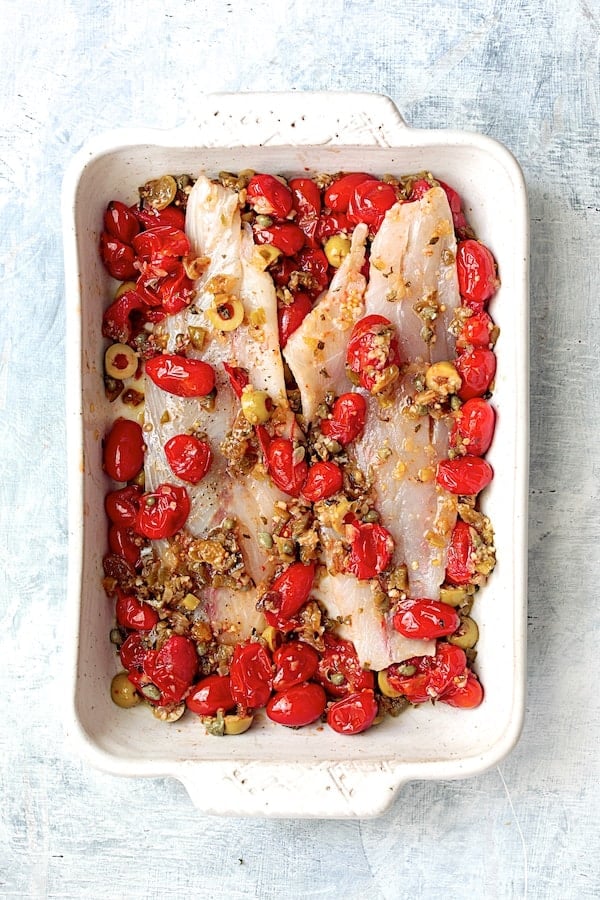 Photo of fish with tomato/olive combination before in white baking dish before being baked.