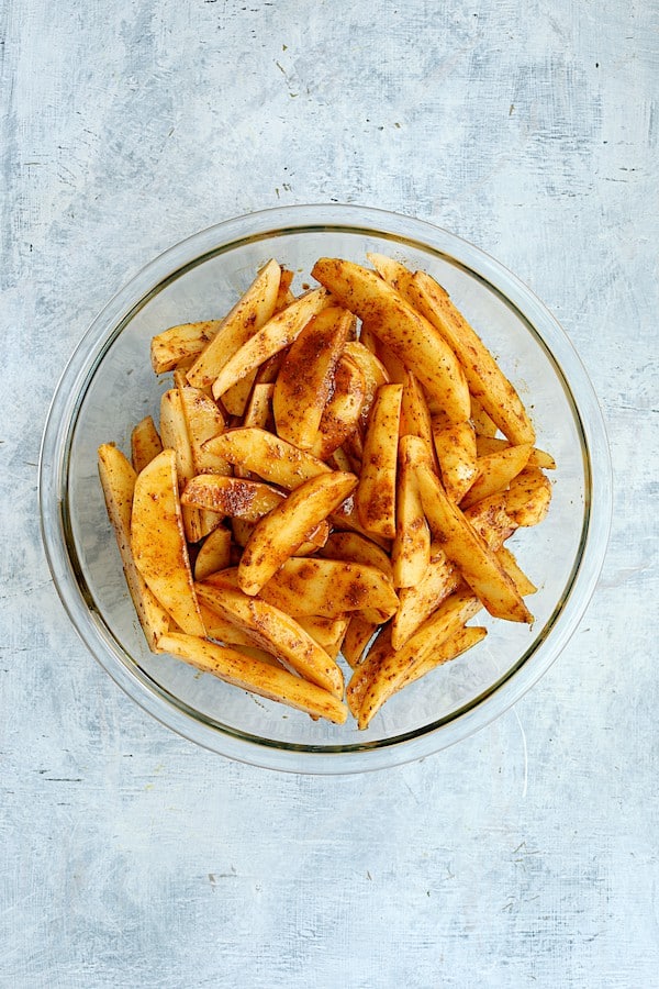 Photo of uncooked potato wedges tossed with oil and Old Bay Seasoning in glass bowl.