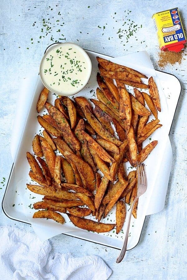 Old Bay Roasted Potato Wedges on white enameled pan with cheese sauce garnished with fresh chives and can of Old Bay.