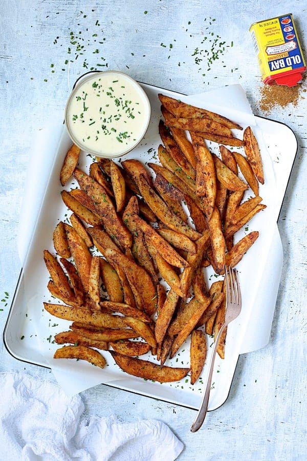 Photo of Old Bay Roasted Potato Wedges with Vermont Cheddar Cheese Sauce on white enameled pan garnished with fresh chives and can of Old Bay.