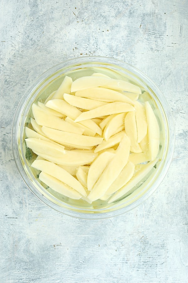 Photo of uncooked potato wedges in water in glass bowl.
