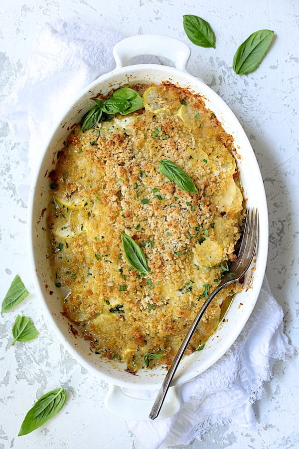 Photo of Yellow Squash and Tomato Gratin with Two Cheeses on white distressed background with white towel garnished with fresh basil.