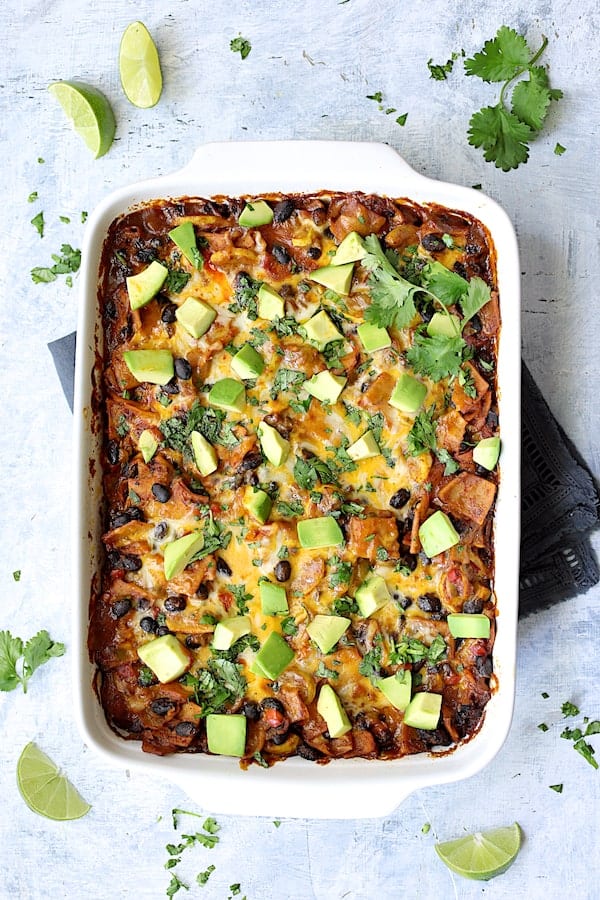 Photo of baked casserole in white baking dish garnished with cilantro, lime wedges and avocado.