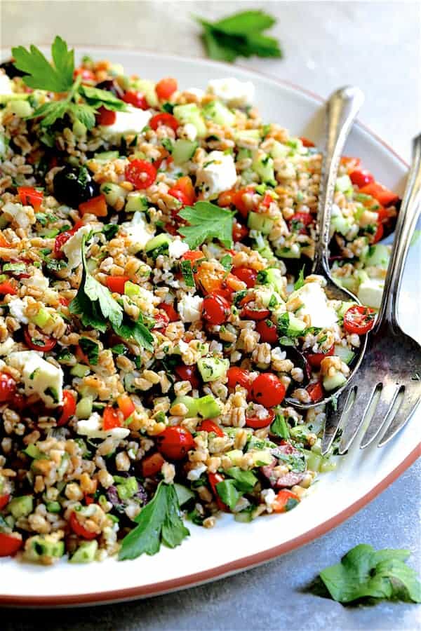 Close-up photo of Mediterranean Farro Salad on white platter with serving utensils.
