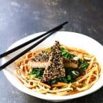 Two pieces of sesame tofu on rice noodles in white bowl with black chopsticks.