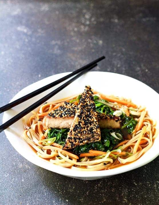 Two pieces of sesame tofu on rice noodles in white bowl with black chopsticks.