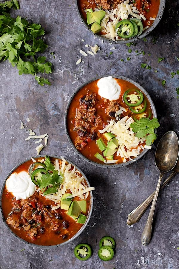 Turkey, Black Bean and Quinoa Chili - Overhead shot three bowls of the chili on gray distressed background garnished with cilantro, jalapeno, cheese and avocado