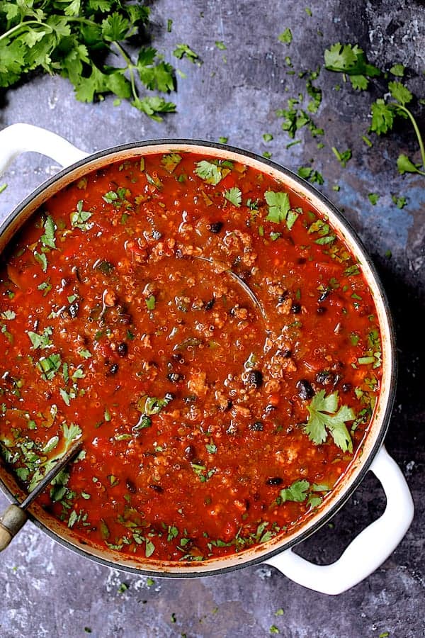 Turkey, Black Bean and Quinoa Chili - Overhead shot of the whole pot of chili in white Dutch oven on gray distressed background garnished with cilantro