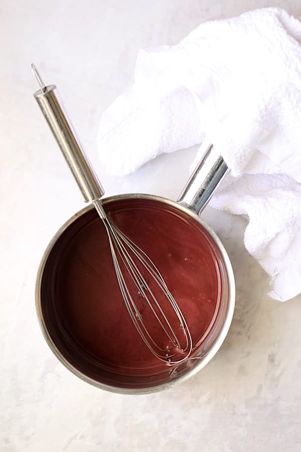 Walnut-Crusted Chicken with Pomegranate Sauce - Overhead shot of sauce in saucepan on white background with whisk and white towel