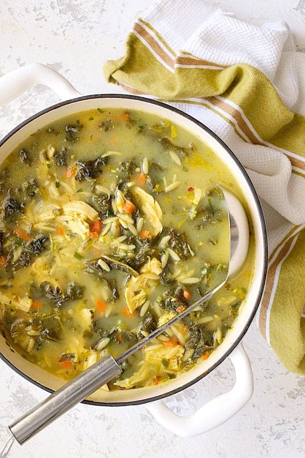 Photo of the whole pot of Cold-Fighting Chicken Soup with Kale and Orzo in white Dutch oven on white background with green striped towel.
